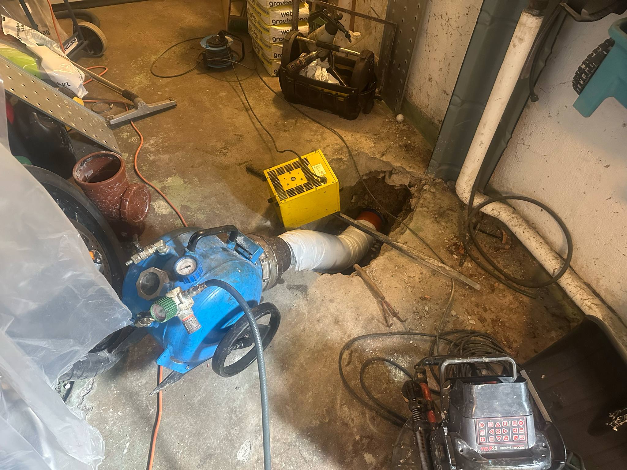Bild på professionell relining och rorsanering i Skarblacka 2025. Ett hål i marken visar rörsystemet under arbete med en blå relining-maskin ansluten. Verktyg, en gul strömbox, verktygslåda och GROWEB-material syns runt omkring i källaren. Optimerad för rorunderhall, rorrenovering och saneringstjanster i Skarblacka.